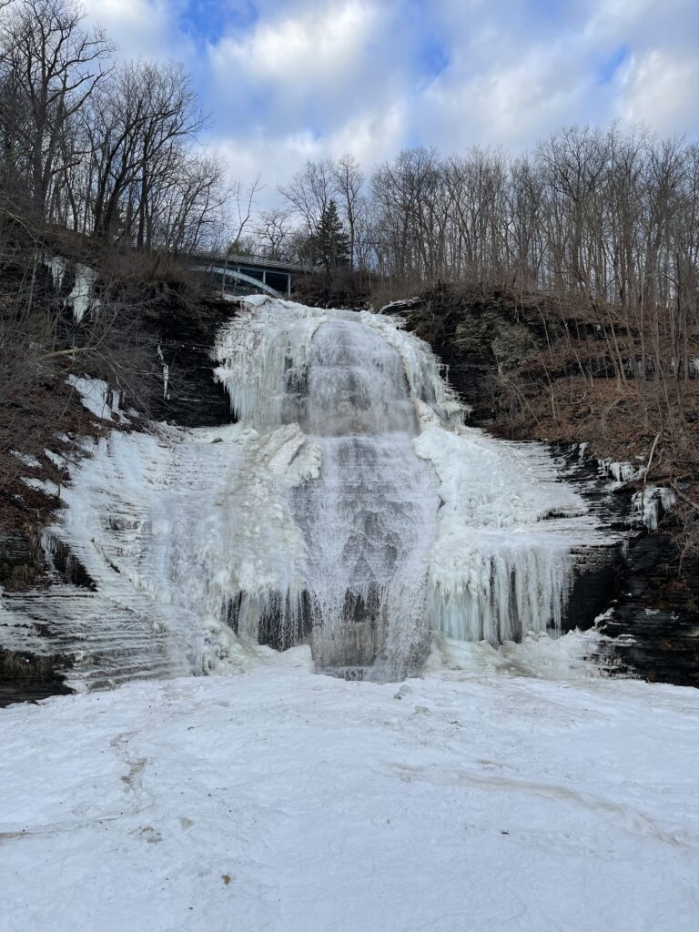 Shequaga Falls partially frozen in winter, icicles on shale ledges and snow on the ground, blue sky and bare trees above, Montour Falls NY