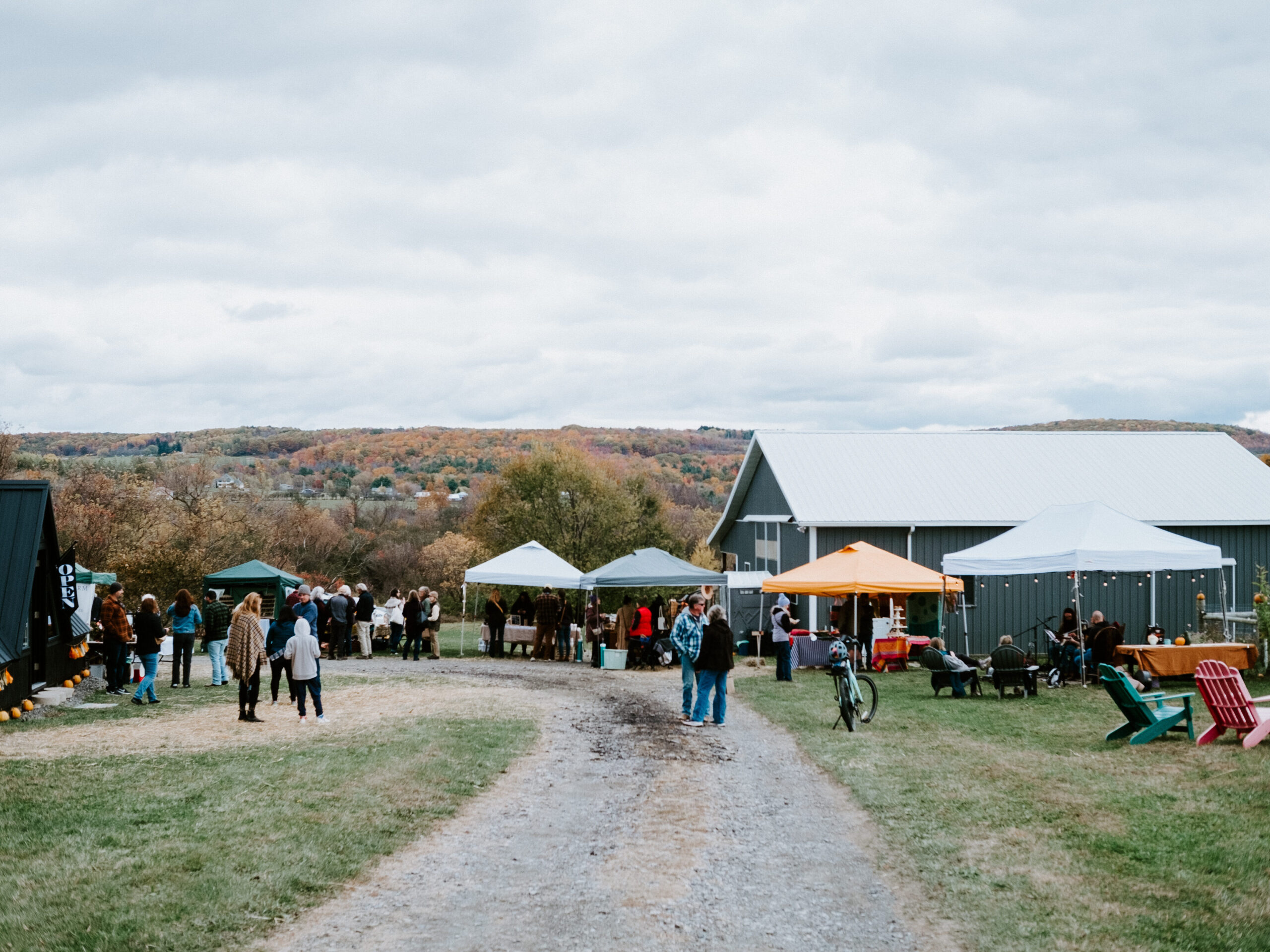 Visitors browsing white and orange vendor tents along a gravel farm lane beside the dark A-frame store and a gray barn, autumn hills behind